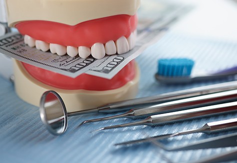 Model of teeth holding money next to dental instruments on table